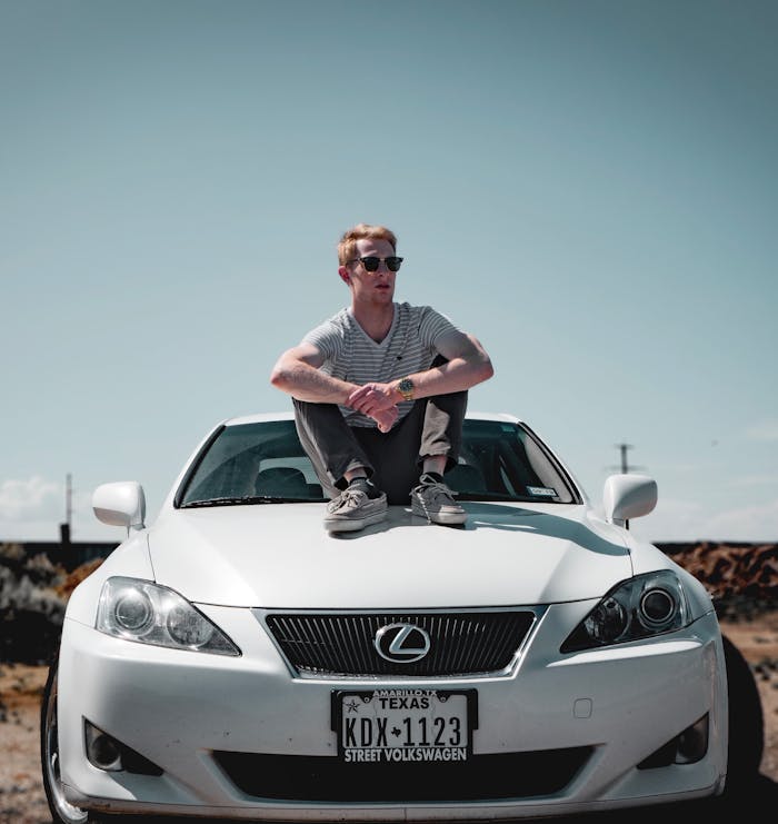 A trendy man sits confidently on a white Lexus, exuding style in a sunlit outdoor setting.